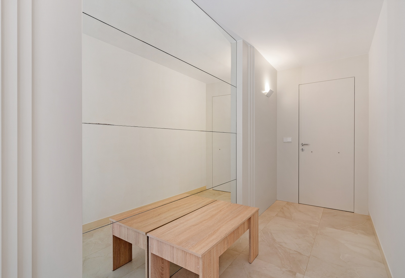 Entrance hallway featuring a large wall mirror, a light wood bench, and cream-toned porcelain floor tiles. Minimalist design with a sleek white door and wall lighting.