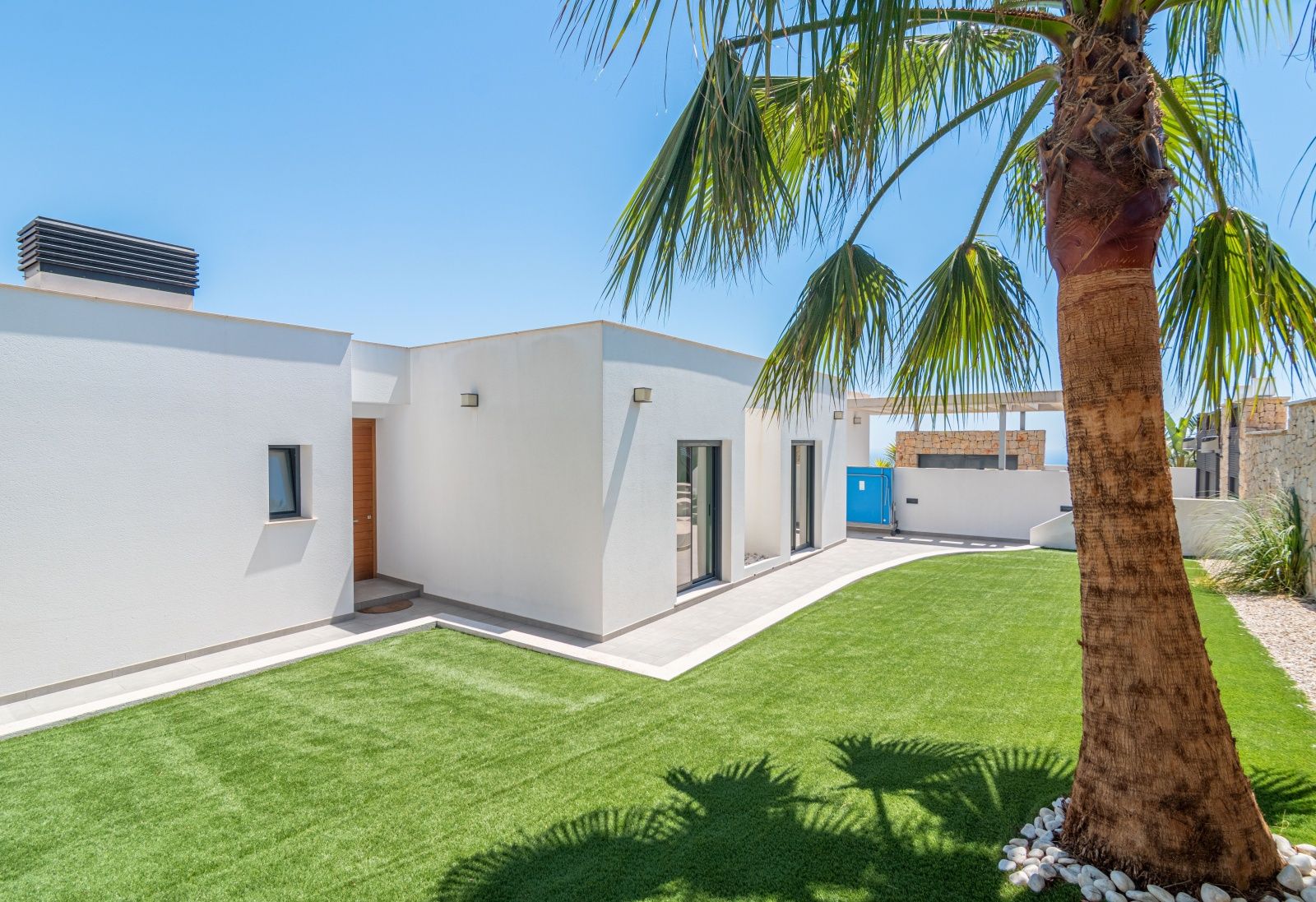 Exterior view of a modern villa with a white facade, artificial lawn, and a palm tree. Features include a wooden entrance door and large windows under a clear blue sky.