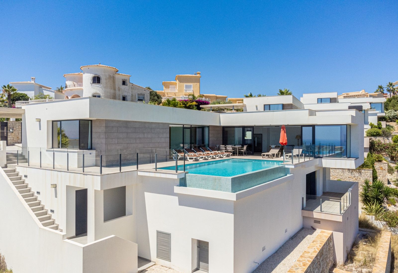Exterior view of the modern villa featuring an infinity pool, spacious sun terrace, and white facade with grey stone accents.
