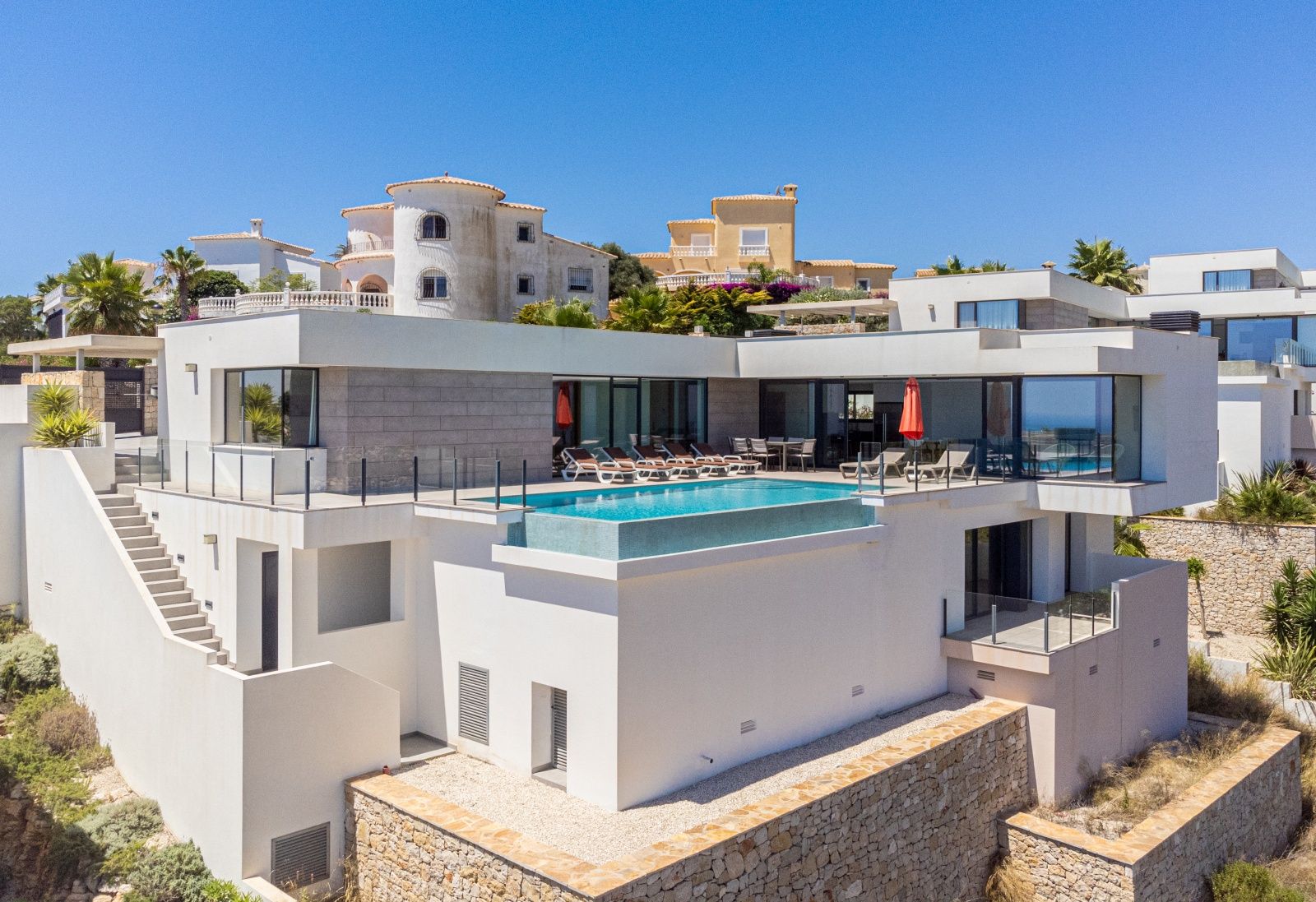 Exterior view of the modern villa featuring an infinity pool, spacious terraces with glass railings, and a white facade with natural stone accents.
