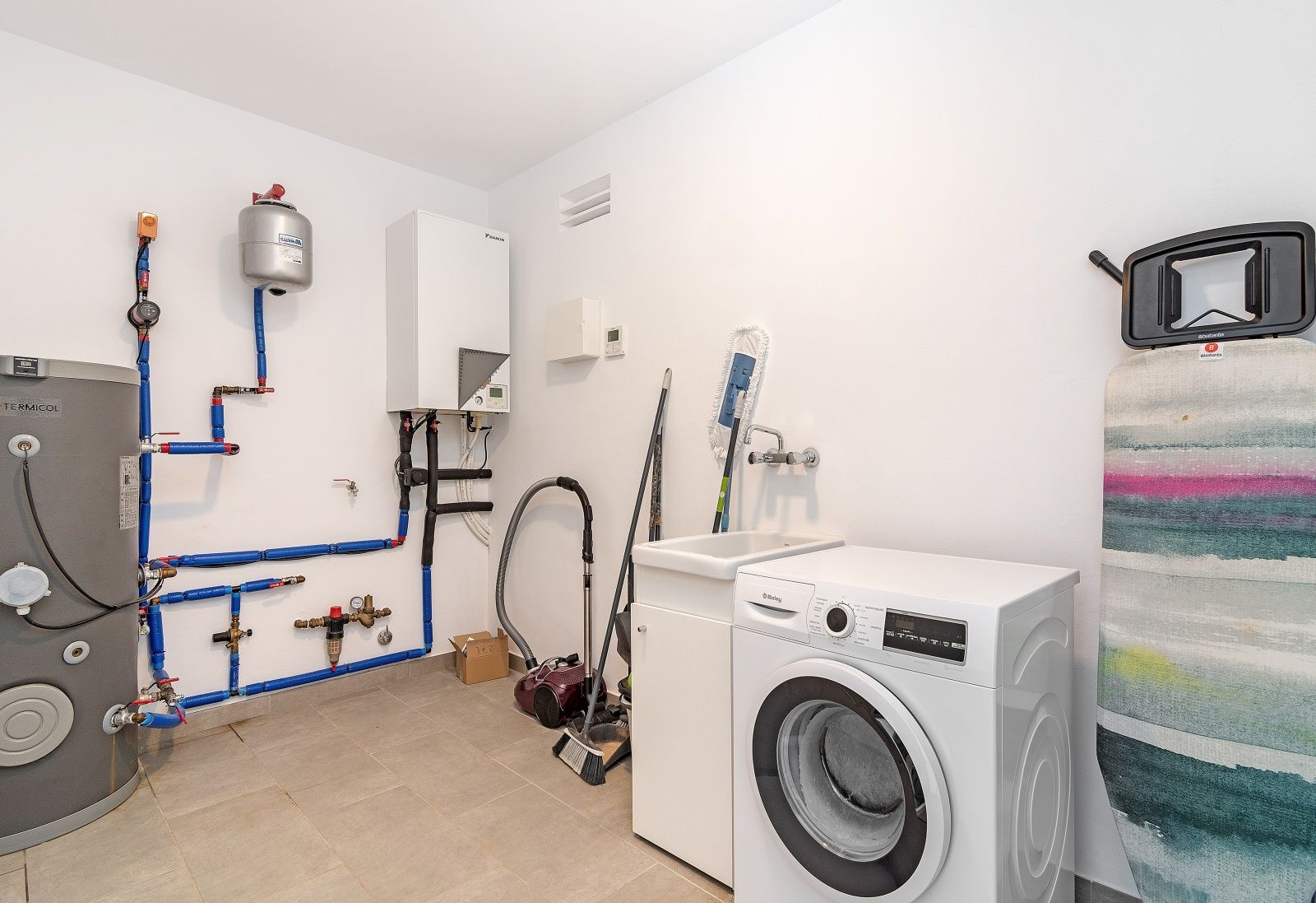 Functional utility room featuring a washing machine, utility sink, and technical installations for heating and water systems with exposed piping.