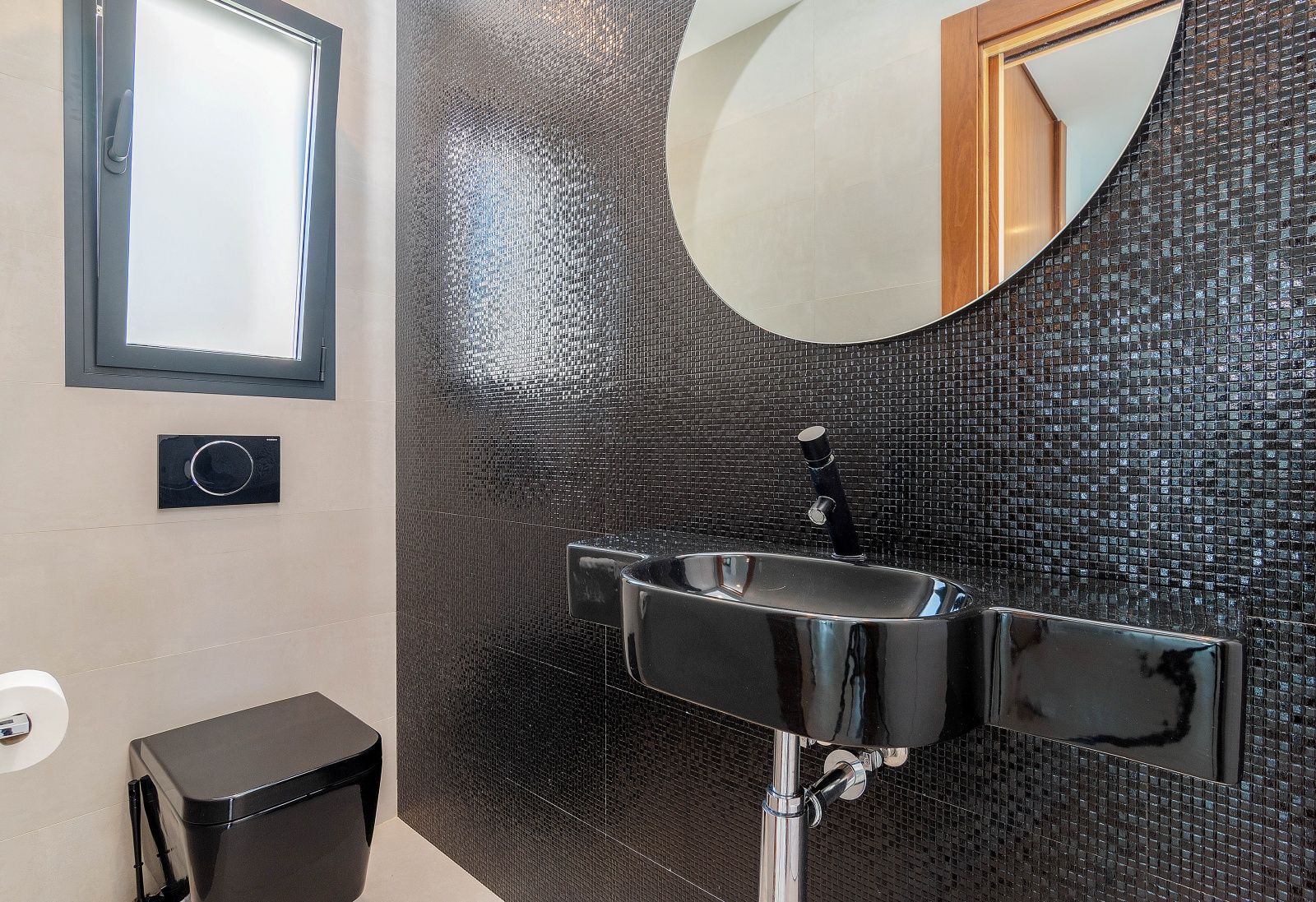Guest bathroom featuring a black mosaic tile accent wall, a matching black ceramic sink, and a wall-hung toilet. A round mirror and frosted window complete the space.