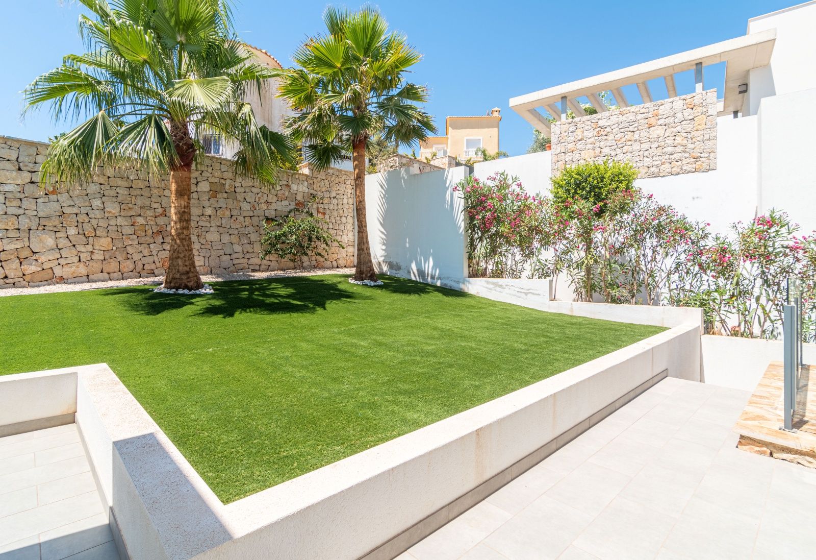 Low-maintenance designer garden featuring artificial turf, palm trees, and natural stone walls integrated with the villa's modern architecture.