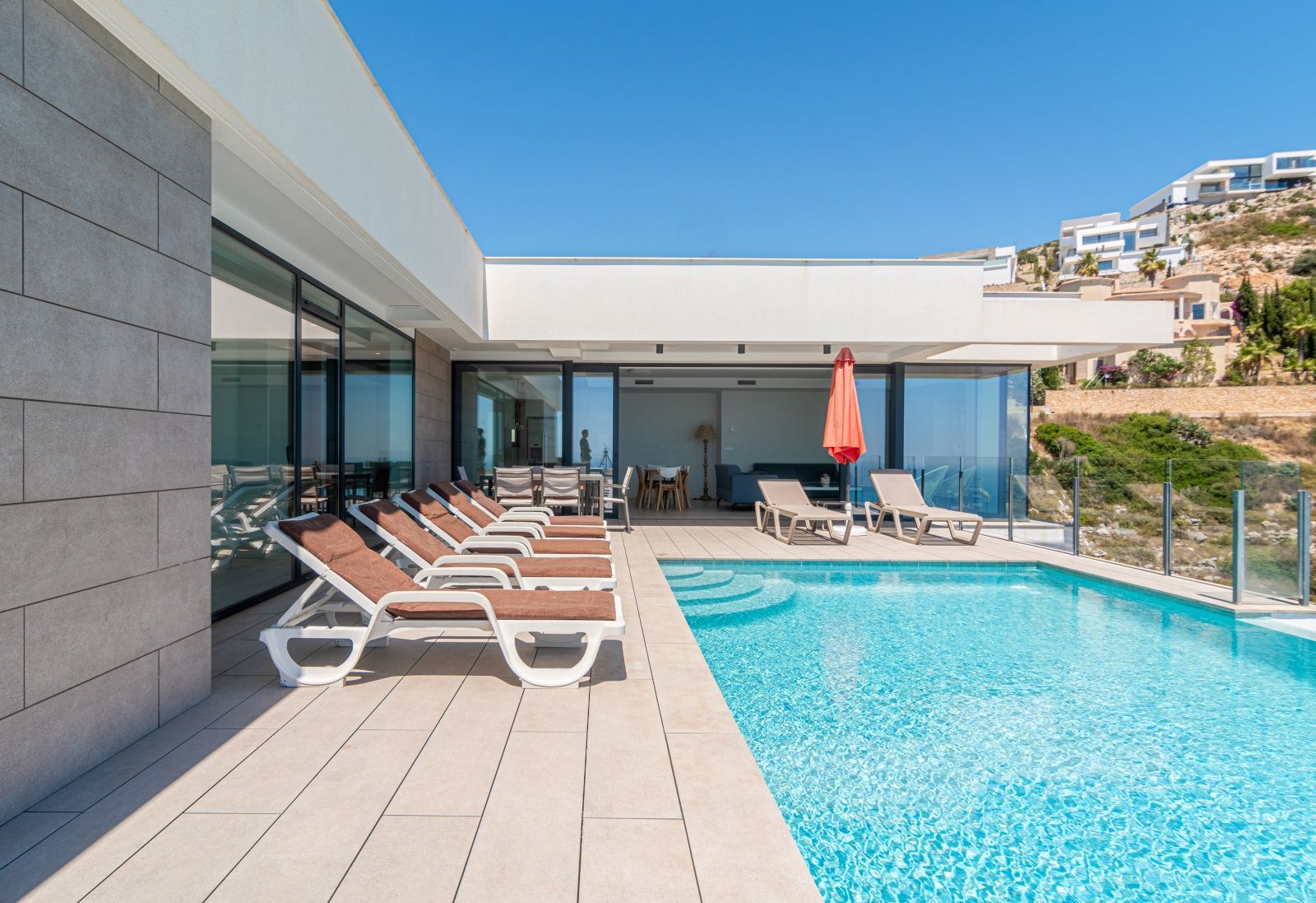Main terrace featuring an infinity pool, sun loungers, and outdoor dining area. The modern architecture includes large glass doors and grey stone wall panels.