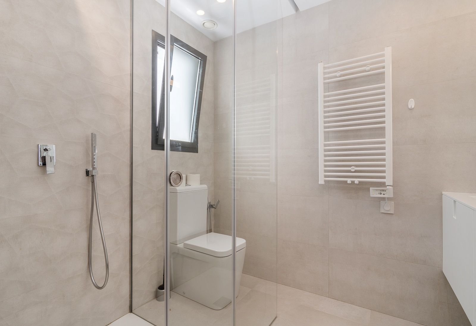 Modern bathroom featuring a glass walk-in shower, textured beige wall tiles, a white heated towel rail, and integrated ceiling spotlights.