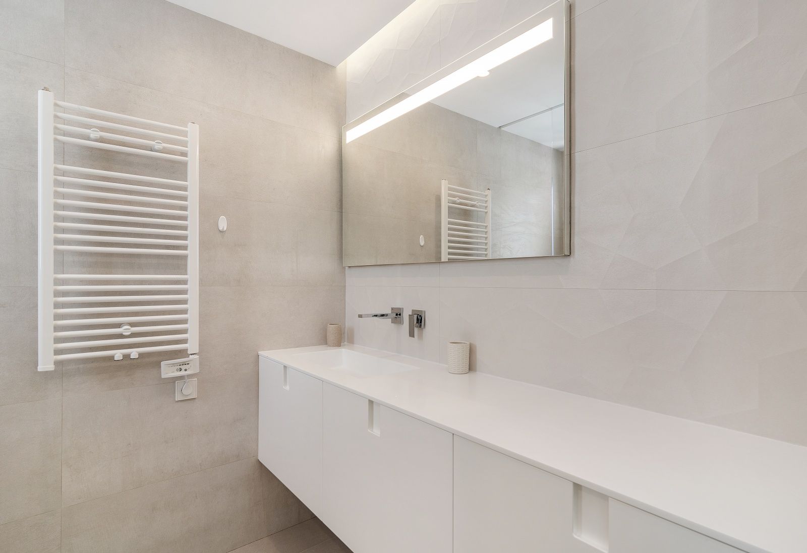 Modern bathroom featuring a white floating vanity, wall-mounted chrome fixtures, a large LED-lit mirror, and a white heated towel rail against textured porcelain walls.
