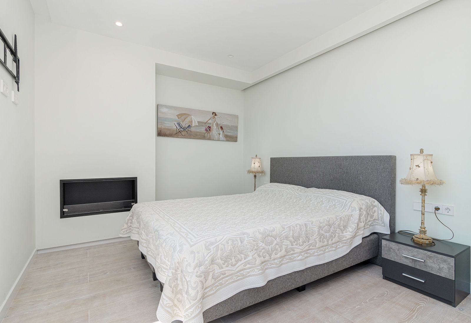 Modern bedroom featuring an integrated wall fireplace, wood-effect porcelain flooring, and a grey upholstered headboard.