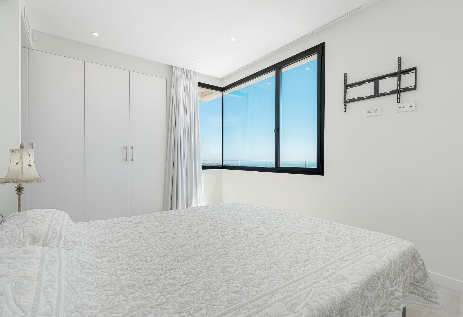 Modern bedroom featuring white built-in wardrobes, a corner window with black frames, and panoramic Mediterranean Sea views.