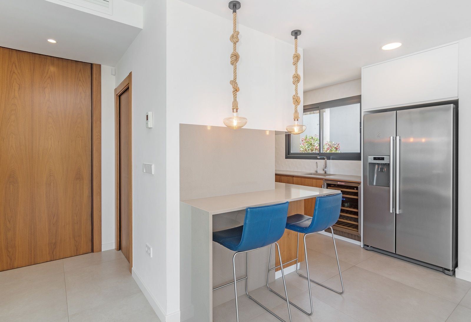 Modern kitchen featuring a breakfast bar with blue stools, rope pendant lights, an integrated wine cooler, and a stainless steel American refrigerator.