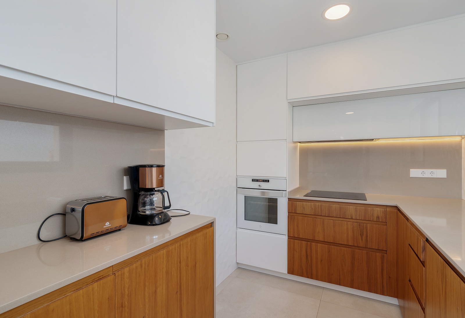 Modern kitchen featuring two-tone white and wood cabinetry, grey quartz countertops, an integrated white oven, and induction cooktop with under-cabinet lighting.