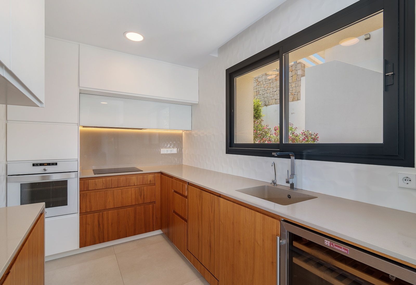 Modern kitchen featuring warm wood and white cabinetry, stone countertops, integrated appliances, a wine cooler, and a large black-framed window.