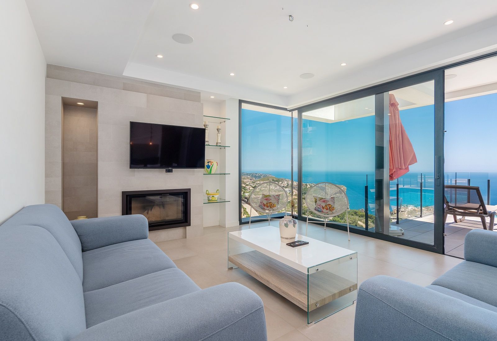 Modern living room featuring blue sofas, a built-in fireplace, and floor-to-ceiling glass doors showcasing panoramic Mediterranean Sea views.