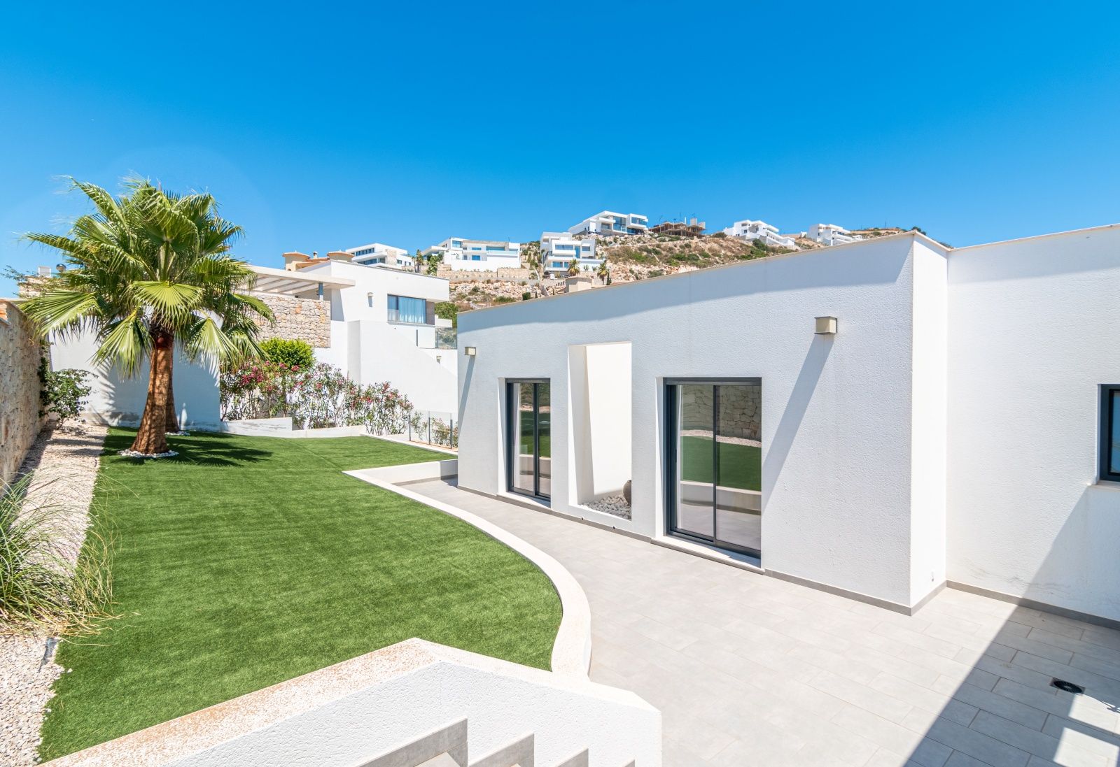 Modern white villa exterior featuring large sliding glass doors, a manicured artificial lawn with a palm tree, and a paved terrace under a clear sky.