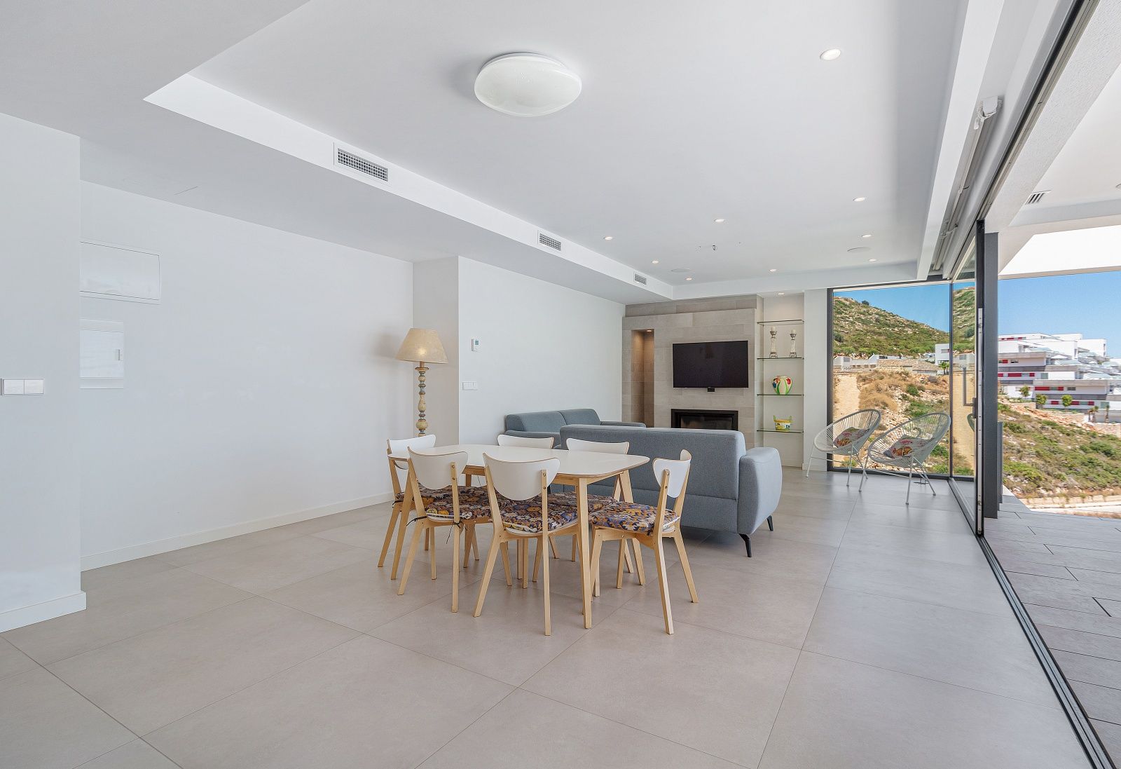 Open-plan living and dining area featuring large format floor tiles, integrated fireplace wall, and floor-to-ceiling glass doors opening to the landscape.