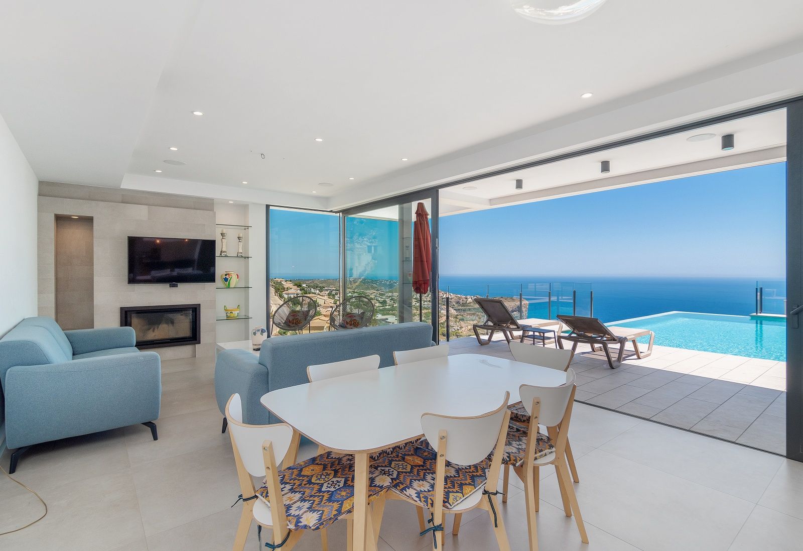 Open-plan living and dining room with a built-in fireplace and large sliding glass doors leading to the infinity pool and panoramic Mediterranean views.