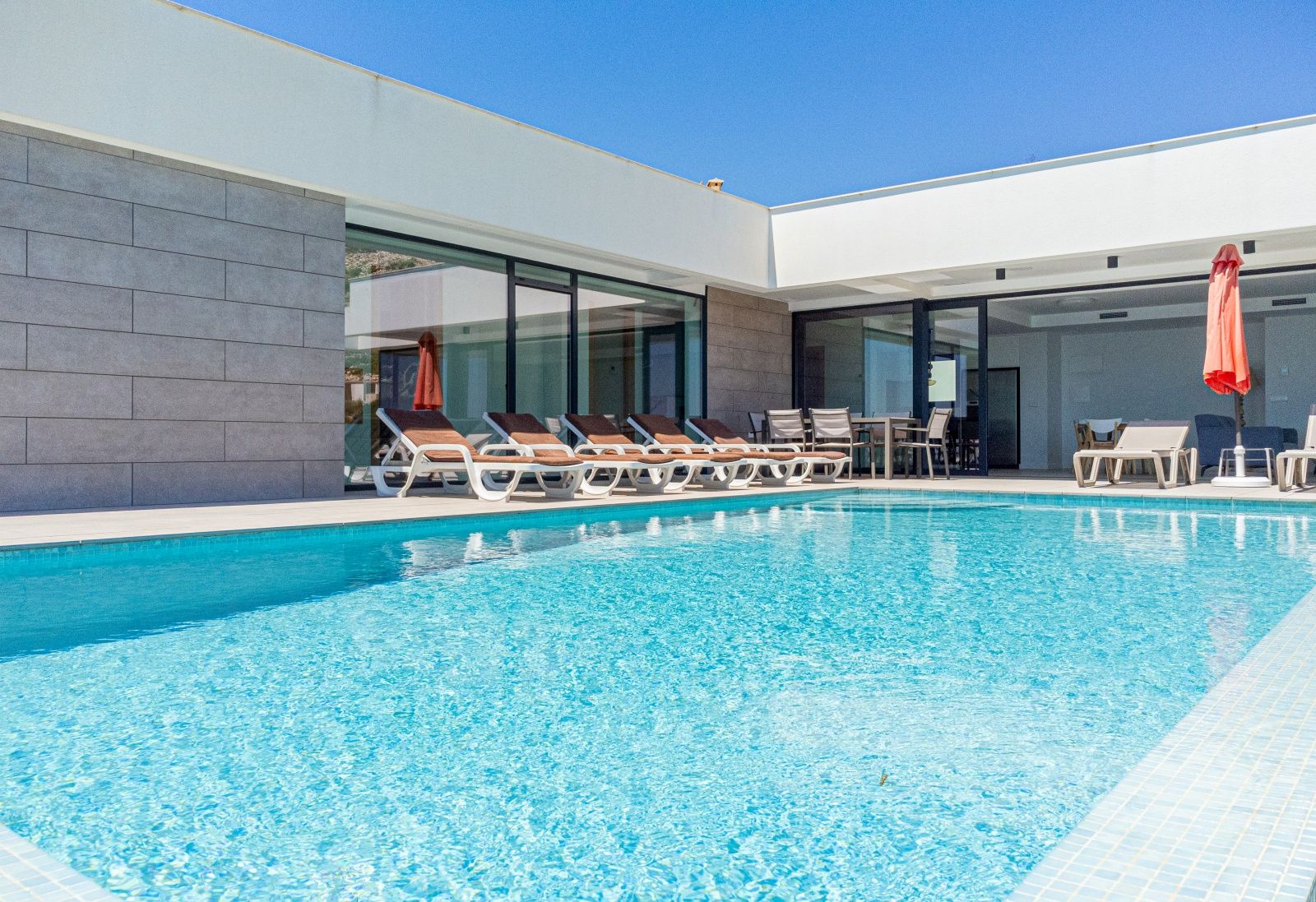 Private pool area featuring a spacious sun deck with loungers, an outdoor dining set, and modern architecture with large glass sliders and stone accents.