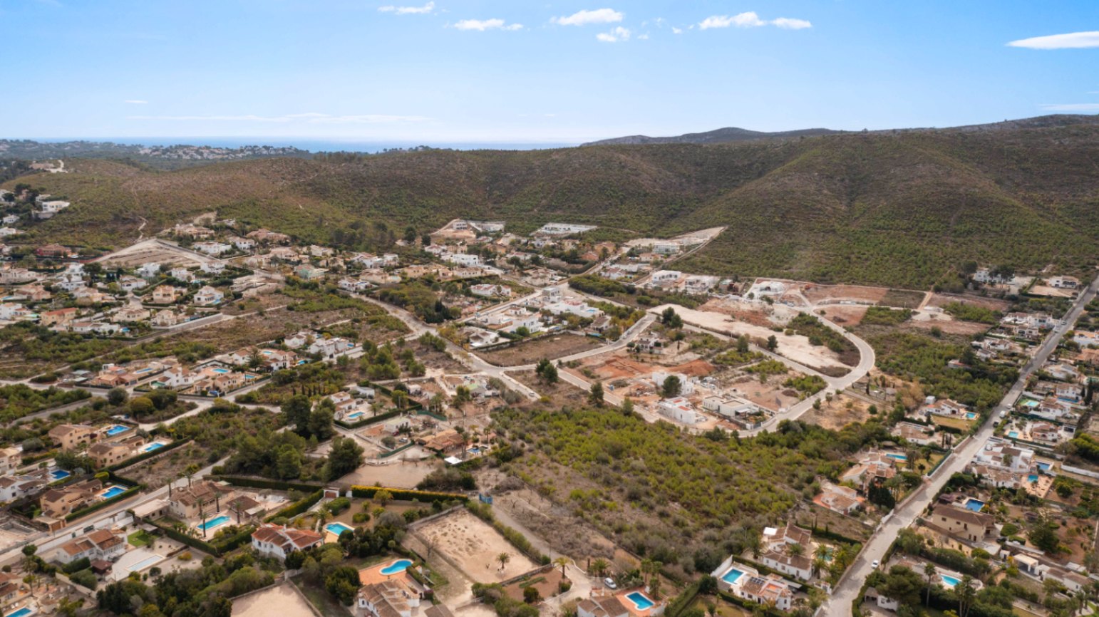 Aerial panoramic view of the Lomas Del Rey residential area in Jávea, showcasing villas with private pools nestled between green hills and the Mediterranean horizon.