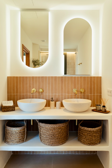 Bathroom featuring double stone vessel sinks, gold wall-mounted faucets, and vertical terracotta tiles. Arched backlit mirrors and wicker storage baskets complete the design.
