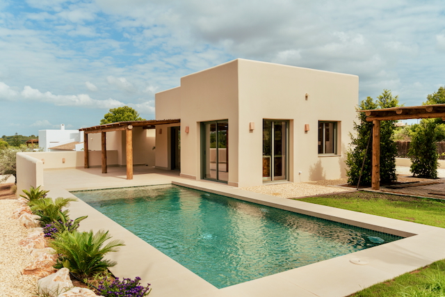Exterior view of the modern villa featuring a rectangular pool, sand-toned facade, and a rustic wooden pergola integrated into the spacious terrace.