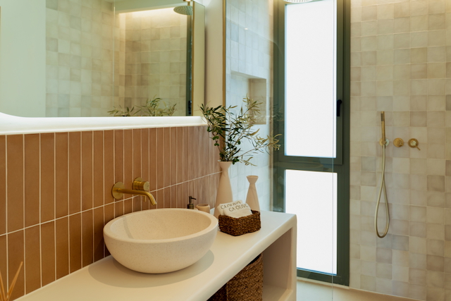 Modern bathroom featuring a stone vessel sink, brushed gold fixtures, and earth-toned vertical tiling. A walk-in shower with a glass partition provides natural light.