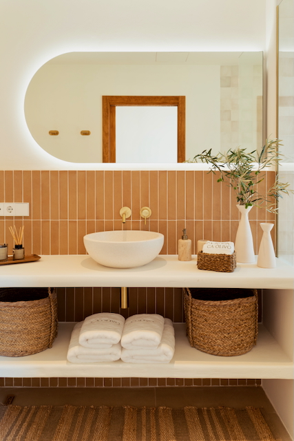 Modern bathroom featuring vertical terracotta tiles, a vessel sink on a white countertop, and gold brass fixtures. A backlit oval mirror complements the open shelving with wicker baskets.