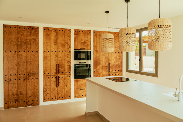 Modern kitchen featuring a white island with induction hob, integrated appliances, and rustic wood cabinetry with stud details. Three wicker pendant lights add a Mediterranean touch.