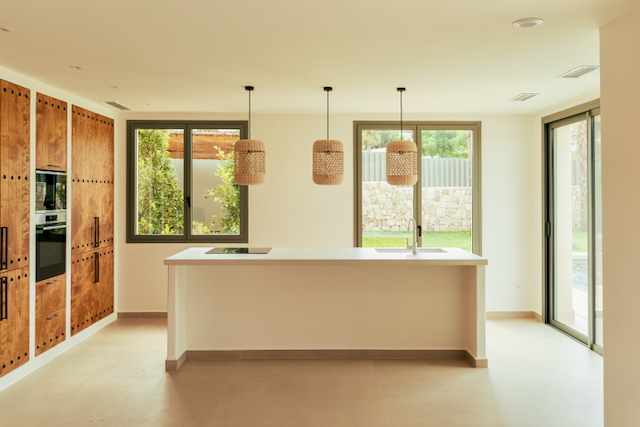 Open-plan kitchen featuring a white island with integrated sink, rattan pendants, and wood cabinetry. Large windows connect the space to the Mediterranean garden.