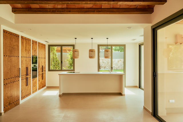 Open-plan kitchen featuring a white minimalist island, rustic wood cabinetry with stud details, and traditional beamed ceilings. Large windows provide abundant natural light.