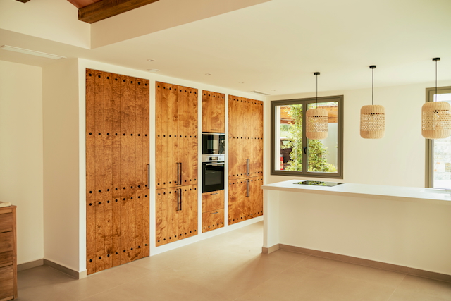 Open-plan kitchen featuring rustic wood cabinetry with studded details and integrated appliances. A white island with induction hob is lit by three woven pendant lamps.