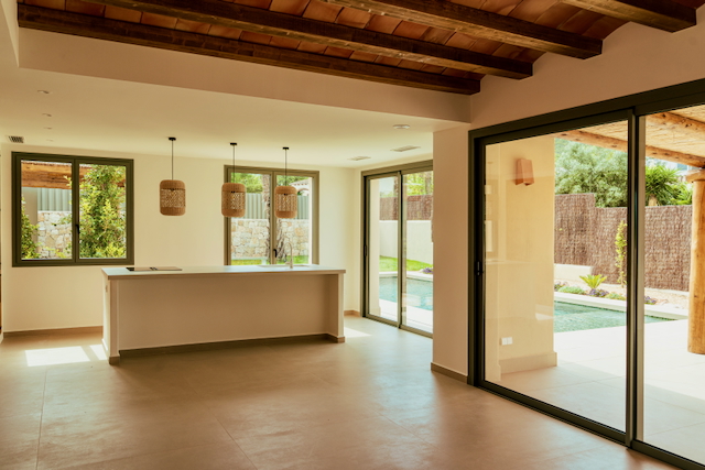 Open-plan living area featuring exposed wooden beams, a kitchen island with woven pendant lights, and large sliding doors opening to the private pool.
