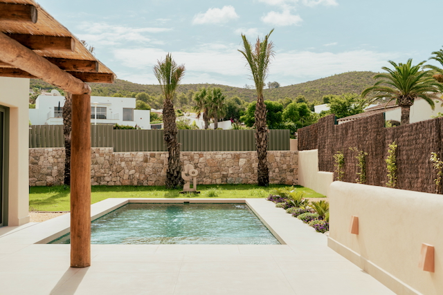 Rectangular swimming pool set in a porcelain terrace, featuring natural stone walls, a Mediterranean garden with palms, and a rustic wooden beam porch.