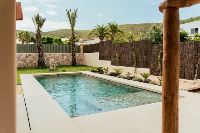 Rectangular swimming pool with light porcelain tiling, surrounded by a landscaped garden with palm trees, stone walls, and scenic hill views.