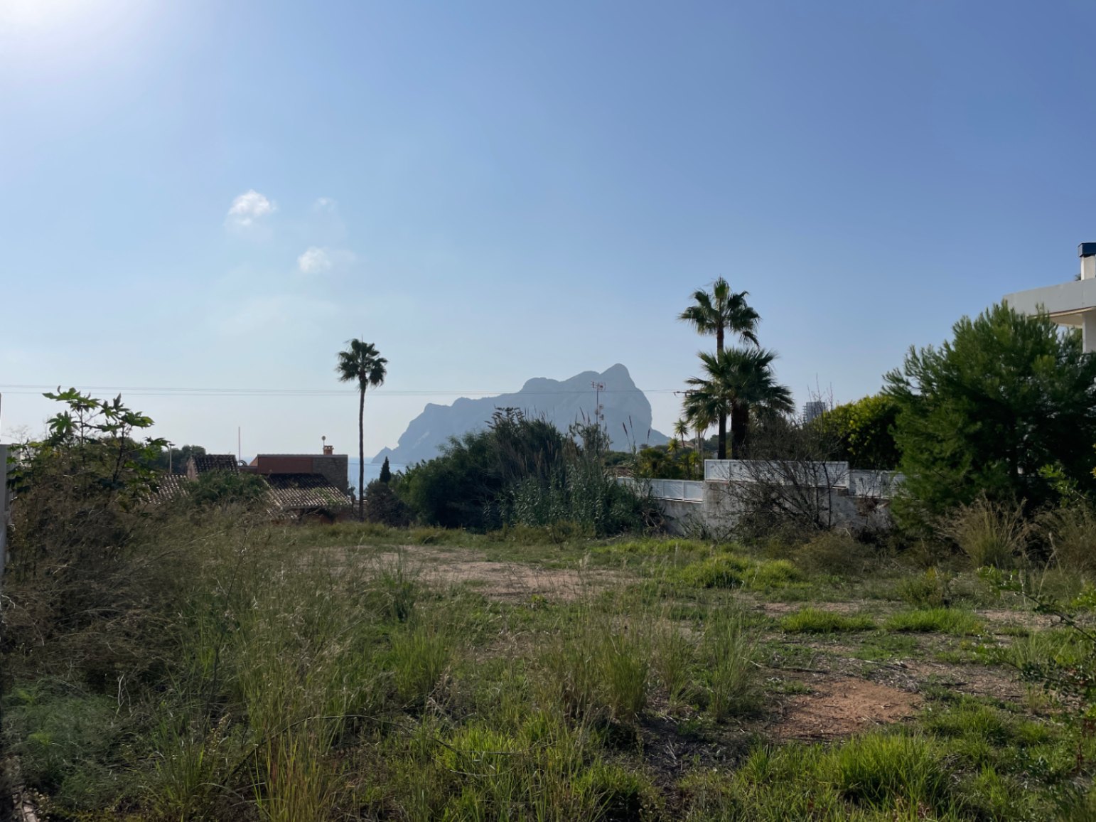 Level building plot in Bassetes featuring direct views of the Peñón de Ifach, bordered by native shrubs and mature palm trees.