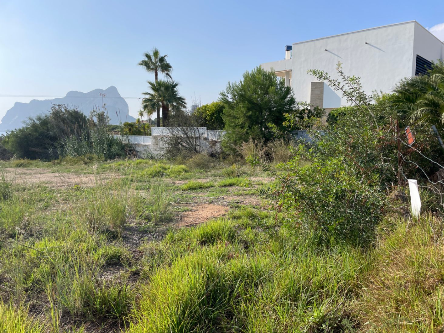 Level plot in Bassetes, Calpe, featuring direct views of the Peñón de Ifach. The land includes natural Mediterranean vegetation and sits adjacent to a contemporary white villa.