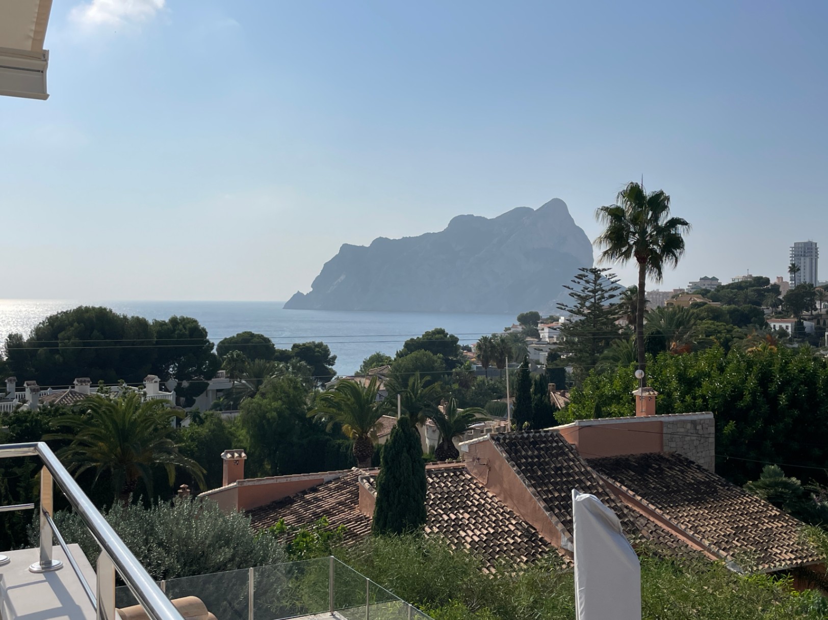 Panoramic view from the terrace overlooking the Peñón de Ifach and the Mediterranean Sea, framed by lush palm trees and traditional tiled rooftops.