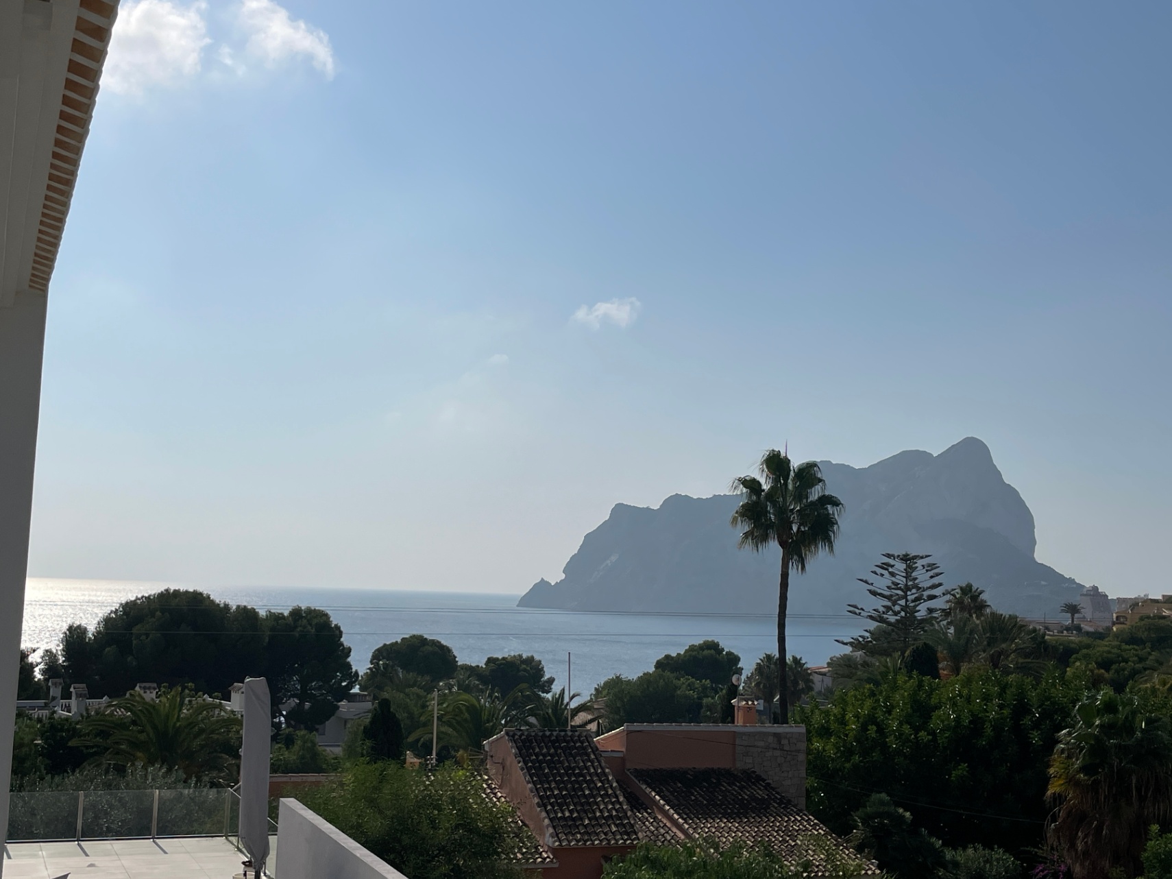 Panoramic views of the Mediterranean Sea and the Peñón de Ifach from an elevated terrace featuring glass railings and surrounding lush vegetation.