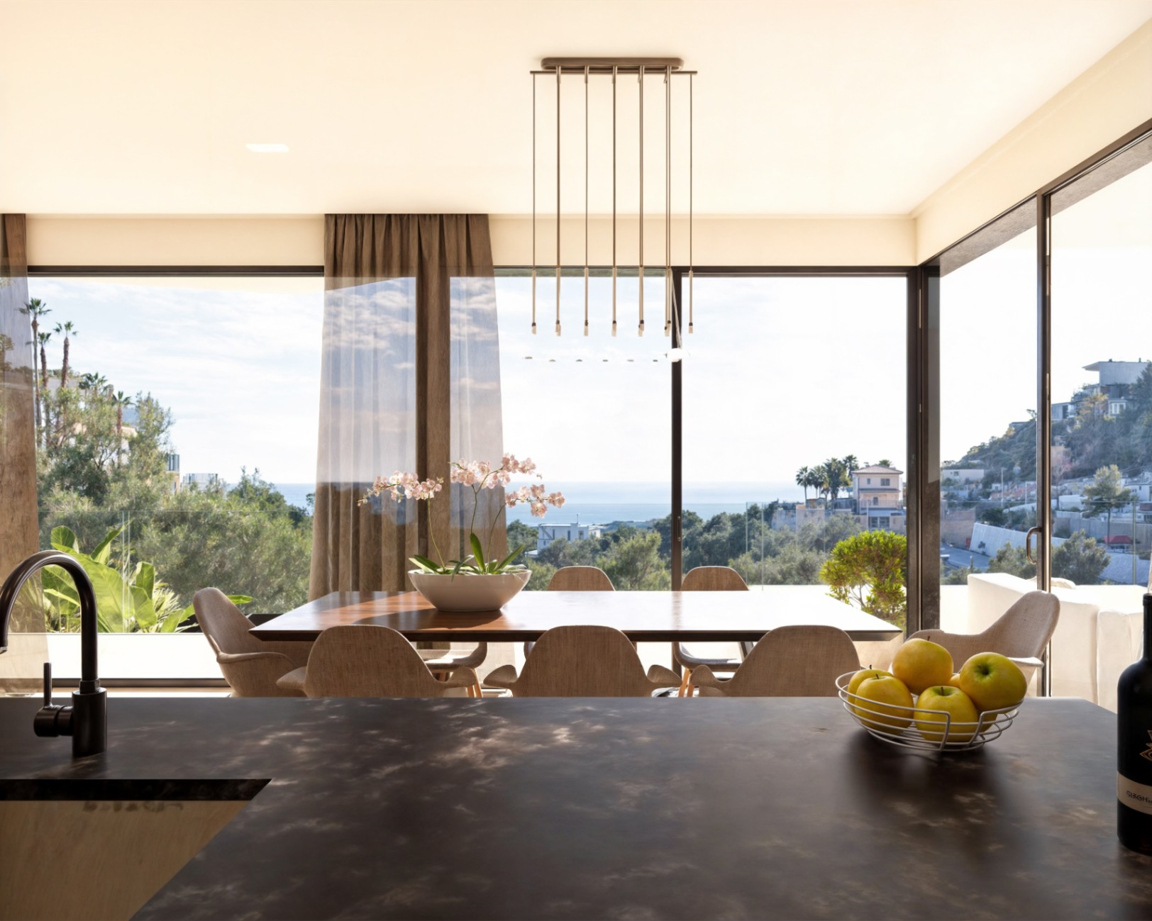 Open-concept dining area featuring a wooden table and floor-to-ceiling windows with Mediterranean views, viewed from a dark textured kitchen island.