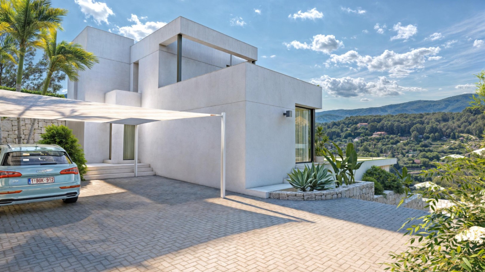 Exterior view of the cubic villa featuring a paved driveway with a shade sail, natural stone walls, and panoramic views of the Sierra de Bernia.