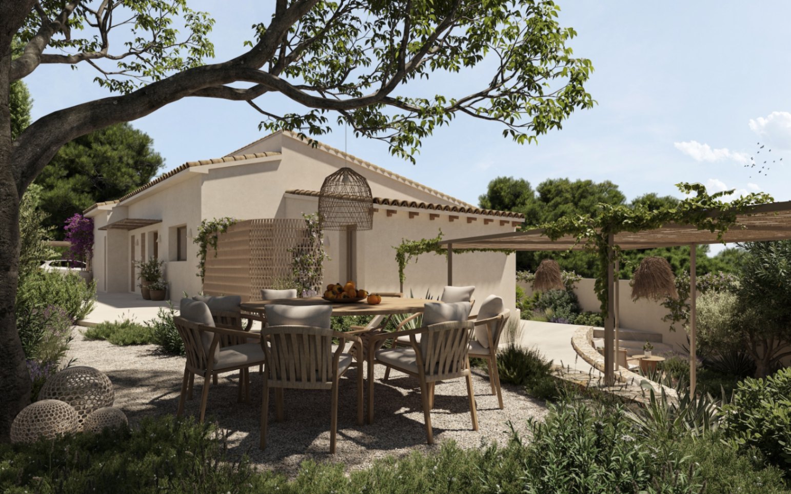 Outdoor dining area shaded by a mature tree, featuring a circular wooden table and woven chairs on a gravel terrace surrounded by Mediterranean gardens.