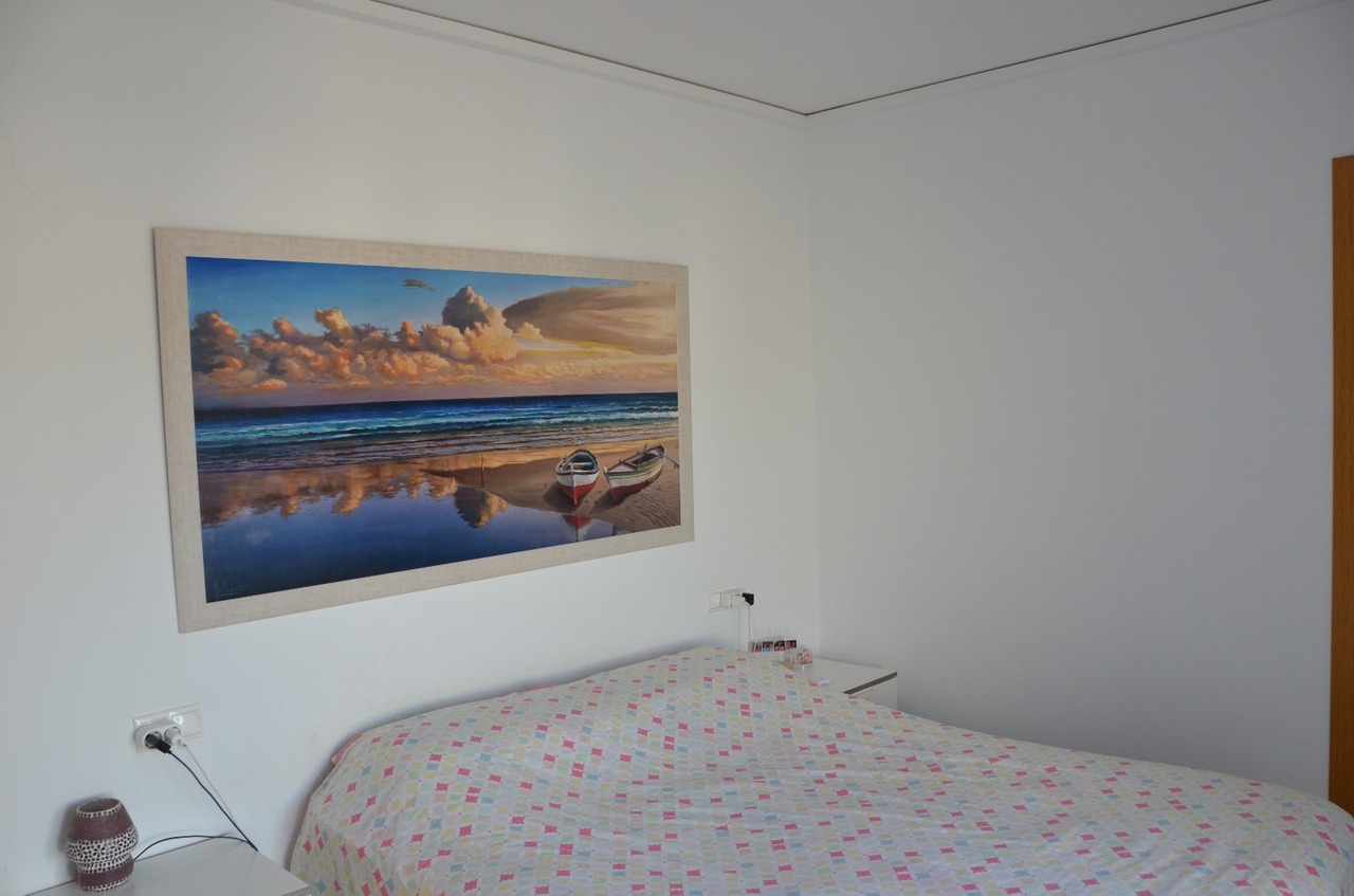 Bedroom featuring a large coastal landscape painting above the bed, white walls, and a bedspread with a colorful geometric pattern.