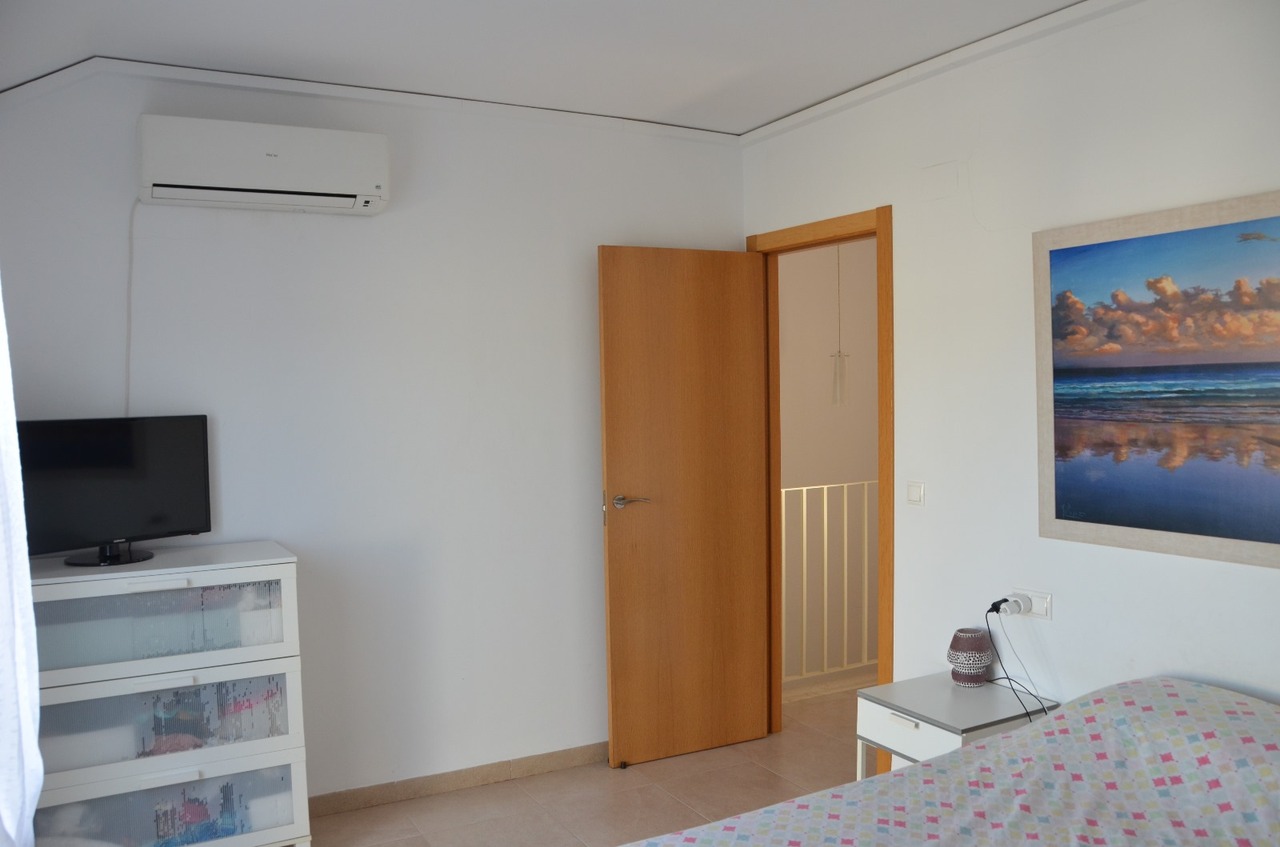 Bedroom featuring a split air conditioning unit, white chest of drawers with a TV, and a wooden door opening to the hallway. Includes tiled flooring and wall art.