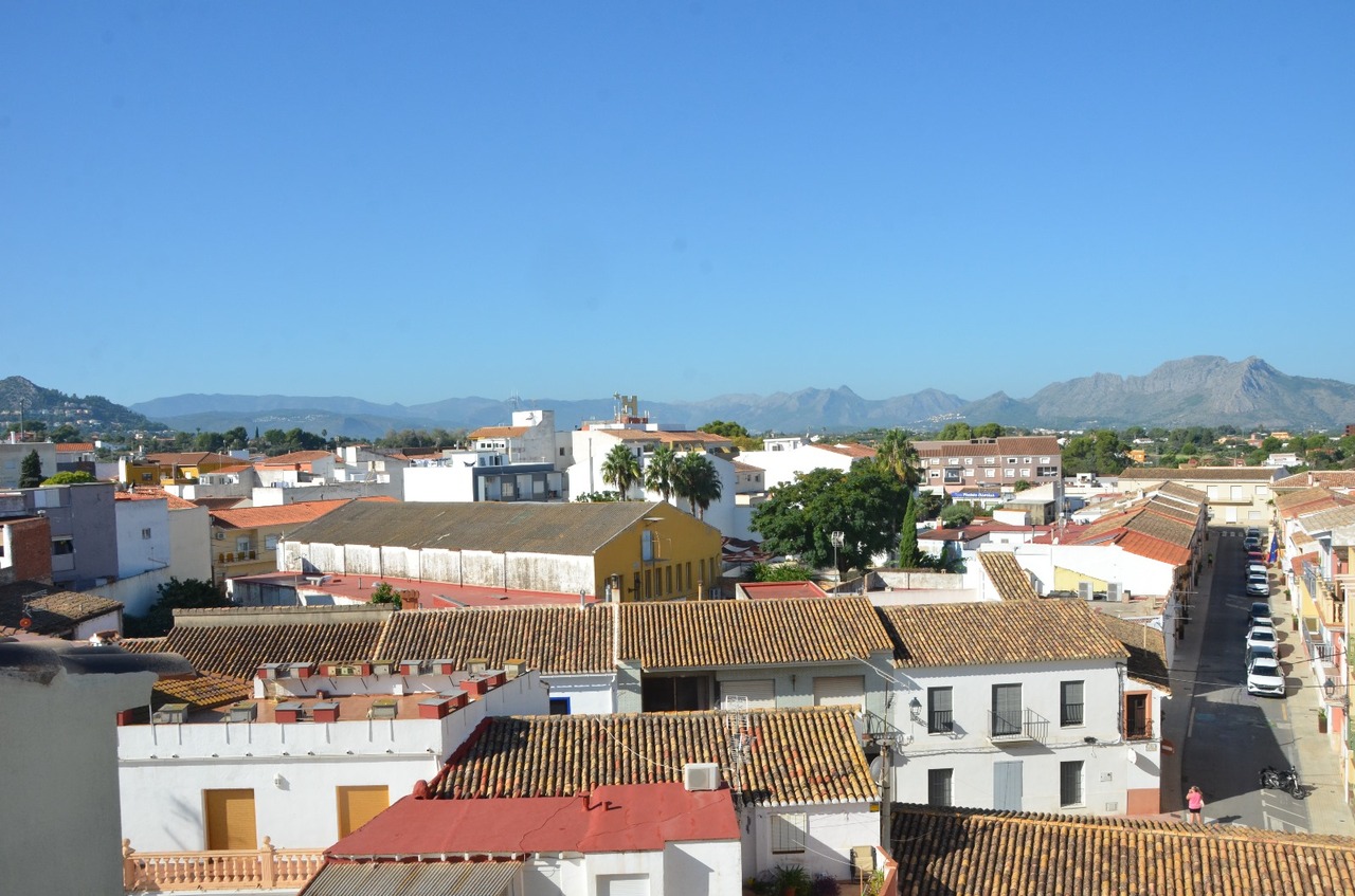 Elevated panoramic view of the town's terracotta rooftops and white-washed buildings, with a clear backdrop of distant mountain ranges under a bright sky.