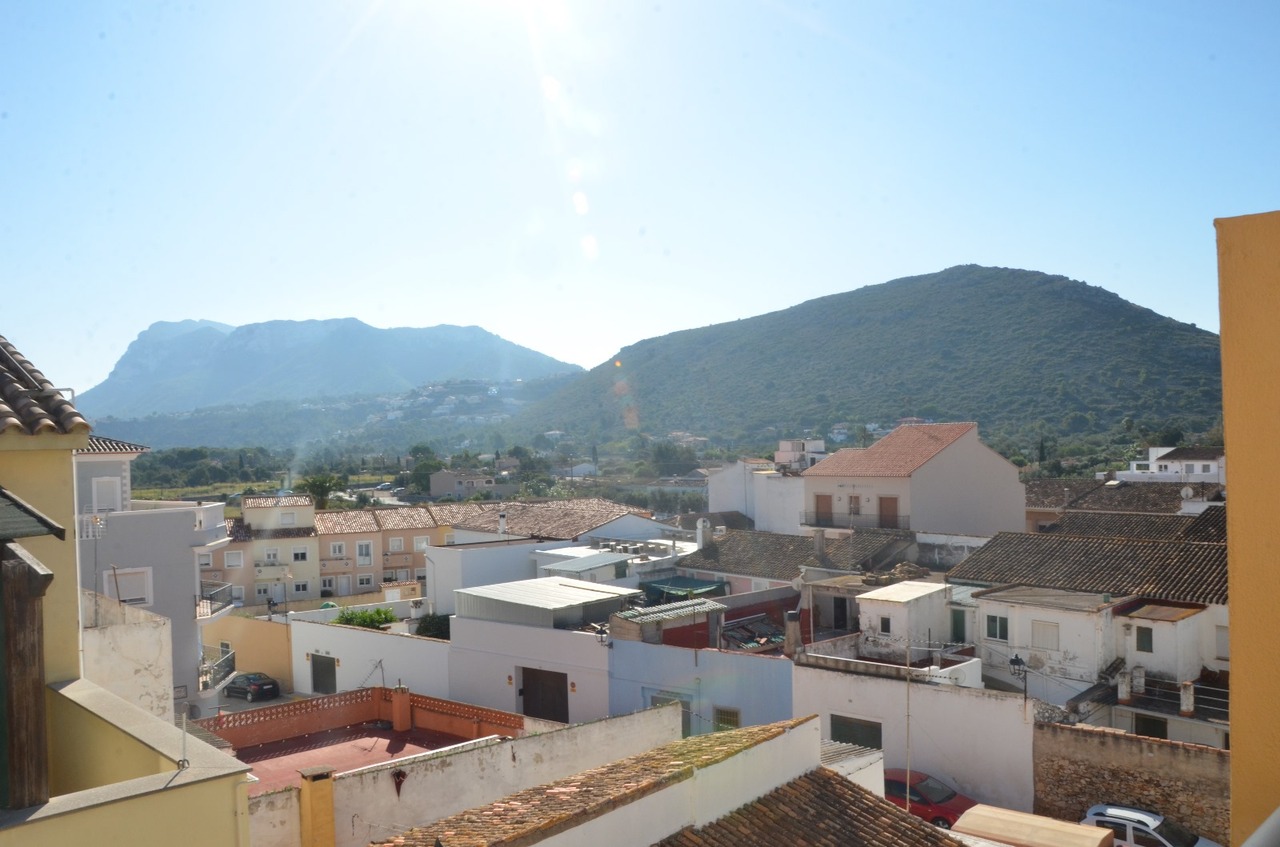 Elevated panoramic view over traditional village rooftops with terracotta tiles, overlooking a lush mountain range under a clear sky.