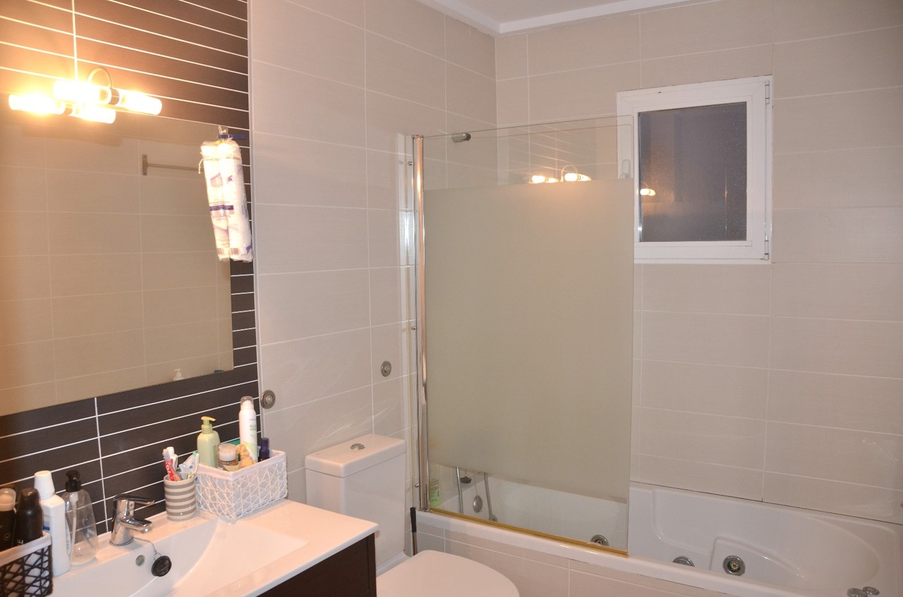 Modern bathroom featuring a white vanity, integrated sink, and a bathtub with a frosted glass partition. Neutral beige tiling is accented by a dark tiled wall behind the mirror.