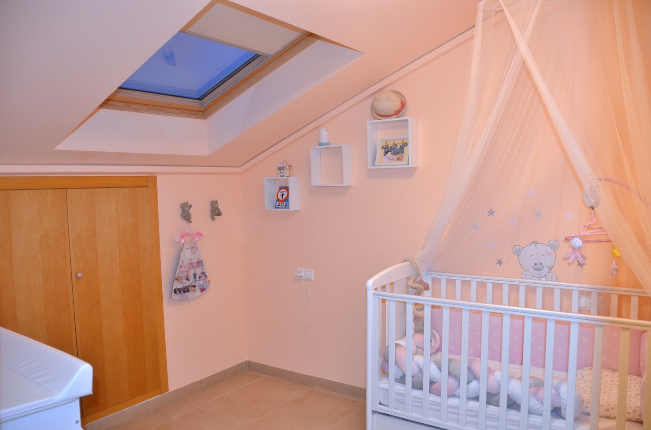 Nursery featuring a sloped ceiling with skylight, white wooden crib with canopy, pink walls, and integrated wooden wardrobe.