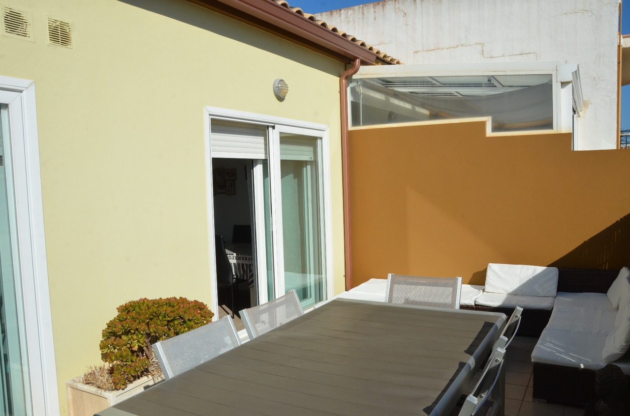 Private outdoor terrace featuring a large dining table with mesh chairs and a corner lounge area with white cushions set against a brown privacy wall.