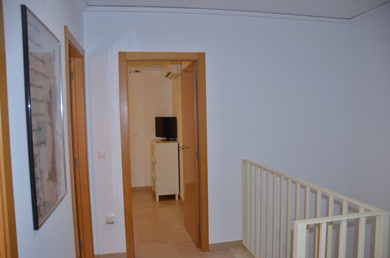 Upper floor landing featuring light wood door frames, white metal staircase railing, and a view into a bedroom with a white dresser and television.