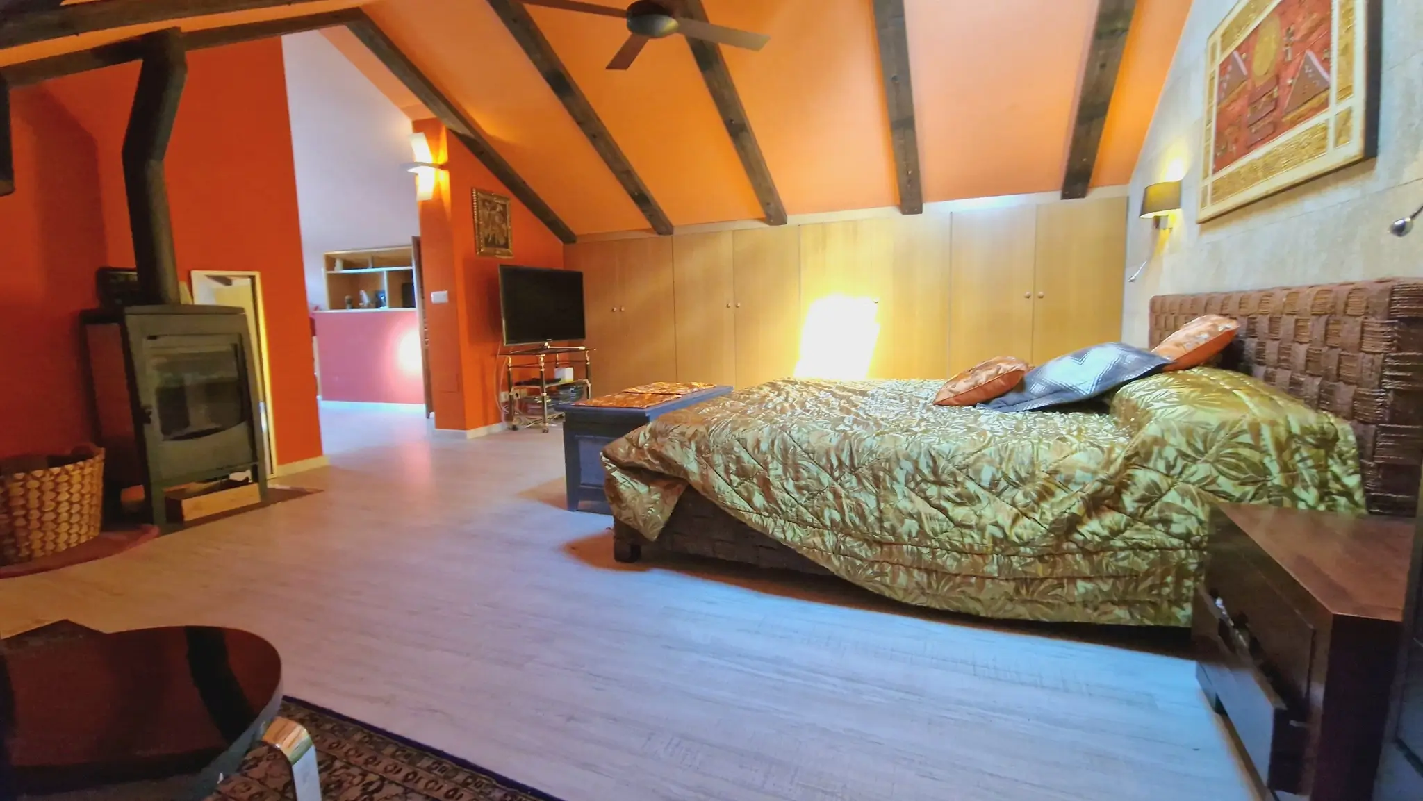 Attic bedroom featuring exposed dark wood beams, a wood-burning stove, and terracotta walls. Includes built-in wardrobes, a ceiling fan, and a woven headboard.