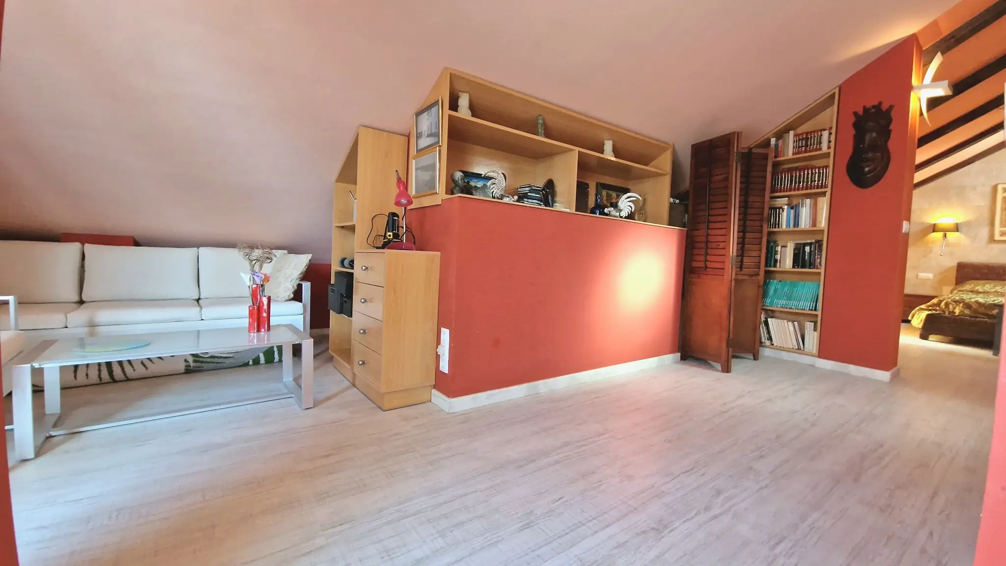 Attic living space featuring terracotta accent walls, integrated wooden shelving, and light-colored laminate flooring under a sloped ceiling.