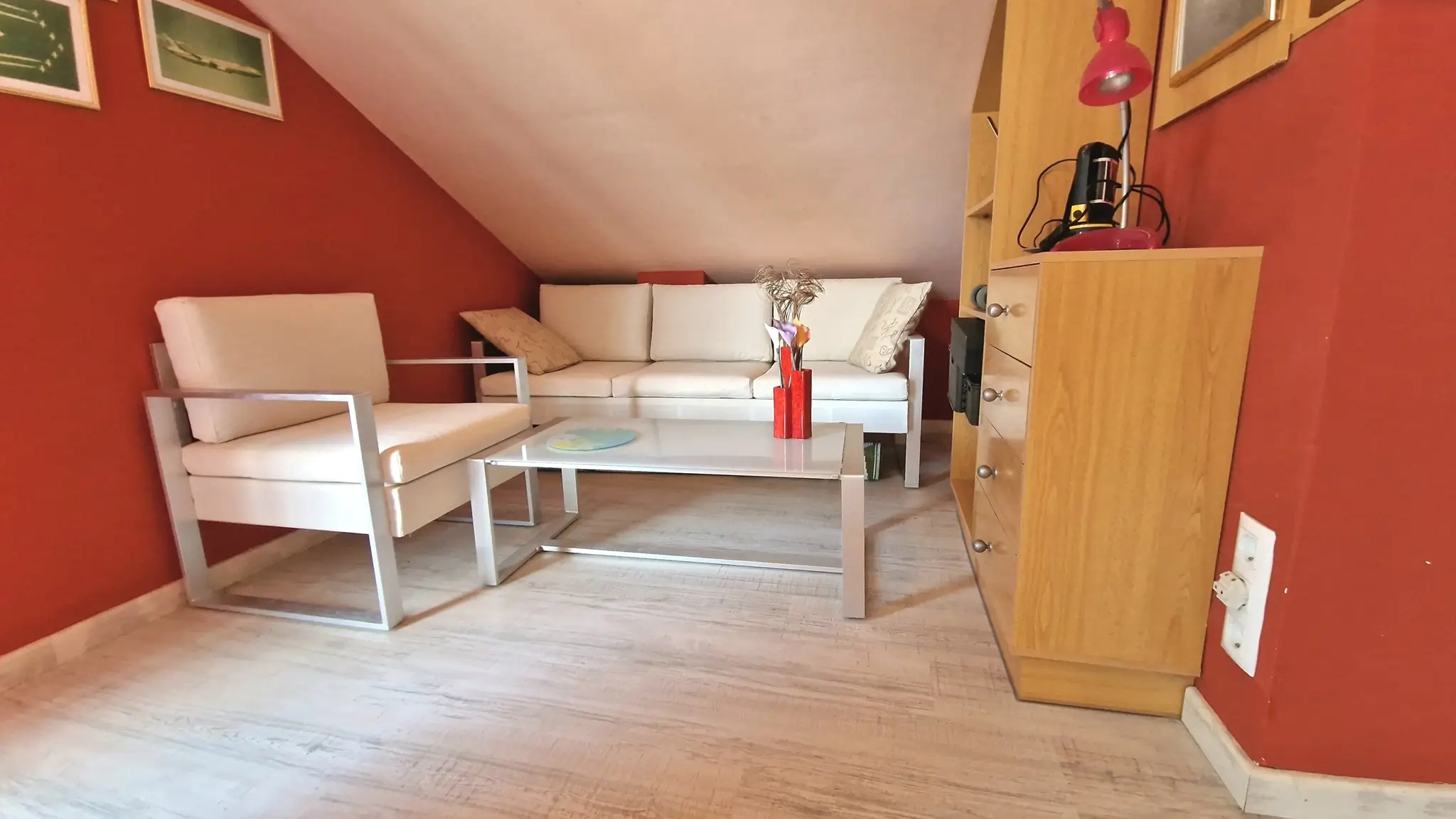 Attic lounge featuring bold red walls, white metal-framed seating, a glass coffee table, and light wood-effect flooring.